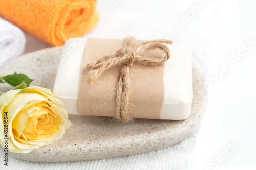 Piece of organic soap wrapped in craft paper on tray with fresh yellow rose and colorful rolled towels.