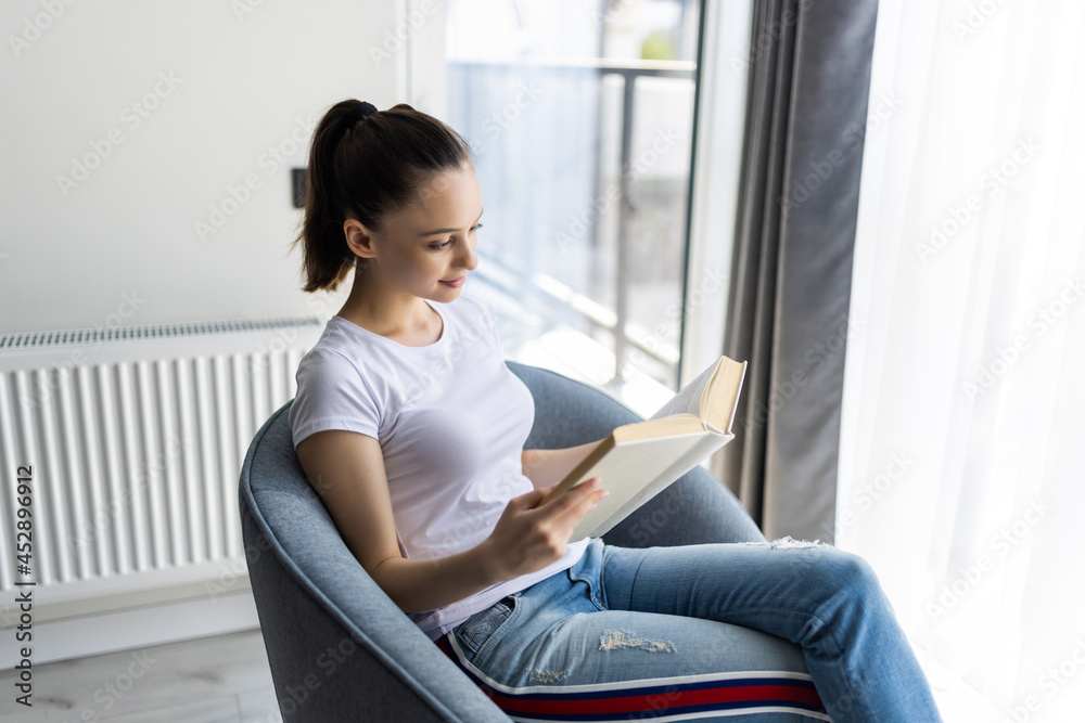 Young woman at home sitting on modern chair in front of window relaxing in her living room reading book Young woman at home sitting on modern chair in front of window relaxing in her living room reading book
