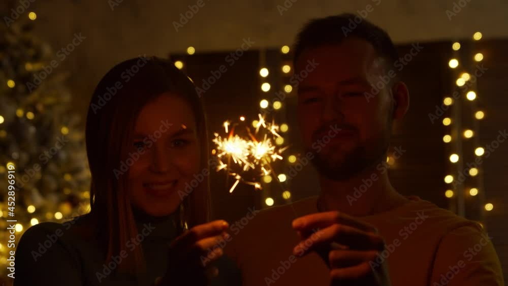 Young loving couple celebrating Christmas at home with a sparklers. Young man and his girlfriend in a home interior with a Christmas tree on the background.
