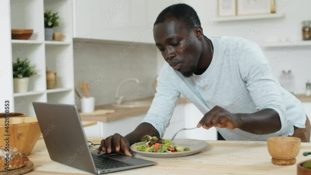 Young Afro-American man leaning on kitchen table, eating salad and surfing the Internet on laptop Young Afro-American man leaning on kitchen table, eating salad and surfing the Internet on laptop