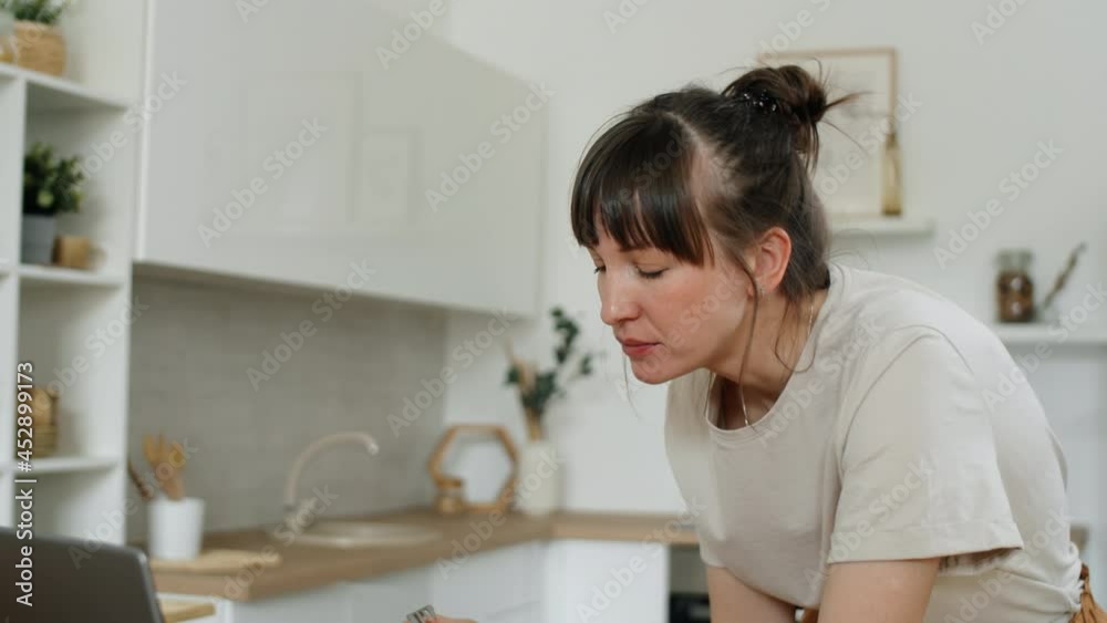 Tilt up shot of young Caucasian woman leaning on kitchen table, eating vegetable salad and browsing the Internet on laptop Tilt up shot of young Caucasian woman leaning on kitchen table, eating vegetable salad and browsing the Internet on laptop