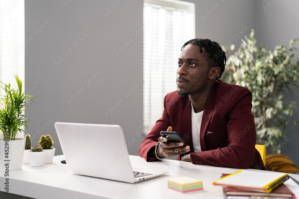 Elegantly dressed dark-skinned businessman sits at white desk in bright spacious office, gray background, laptop, documents, plants, man holds phone in hand, try to remember number, ponders looking up Elegantly dressed dark-skinned businessman sits at white desk in bright spacious office, gray background, laptop, documents, plants, man holds phone in hand, try to remember number, ponders looking up