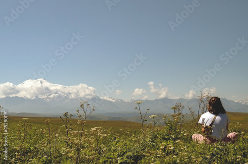 A girl sits in a meadow and looks at a beautiful mountain landscape.