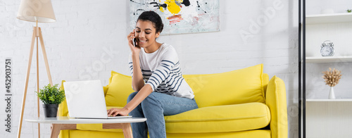 Smiling african american woman talking on smartphone and using laptop in living room, banner