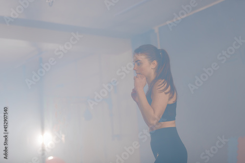Caucasian woman in sportswear with a ponytail, kicking the boxing bag inside a gym with a smokey background