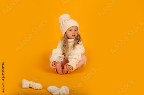 beautiful little blonde girl in a winter knitted white hat sweater and socks new year on a yellow background