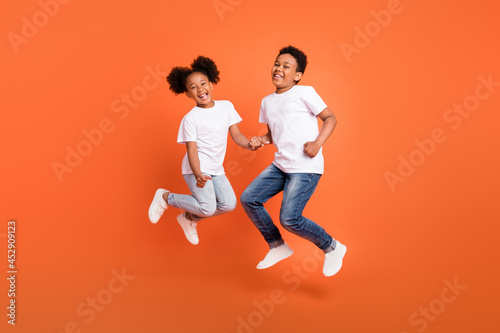 Full length photo of little funky girl boy jump wear white t-shirt jeans sneakers isolated on orange background