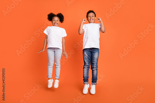 Full length photo of little hooray girl boy jump wear white t-shirt jeans sneakers isolated on orange background