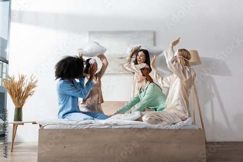 Принти на полотні cheerful young interracial friends in pajamas having pillow fight during slumber