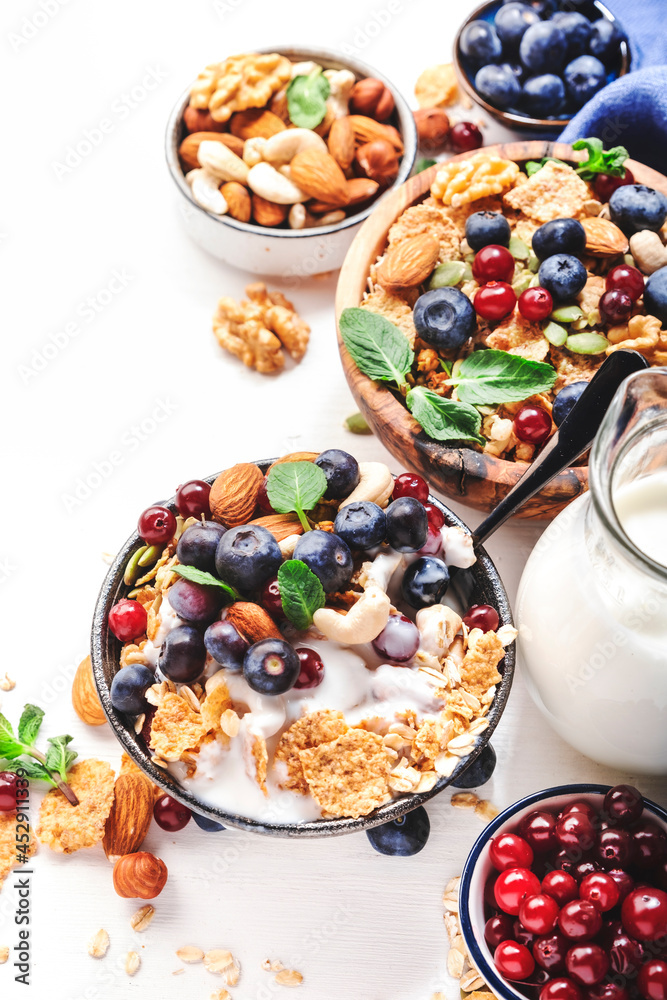 Fototapeta premium Muesli bowl and organic ingredients for healthy breakfast. Granola, nuts, blueberry, cranberry, oatmeal, greek yoghurt, whole grain flakes on white table