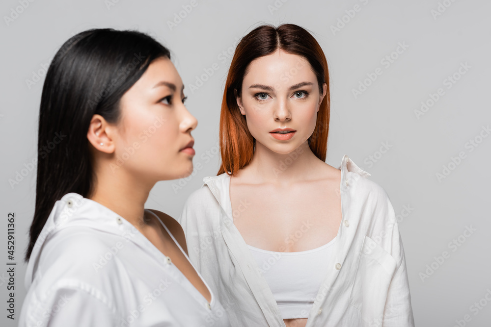 young redhead woman posing near blurred and brunette asian model isolated on grey young redhead woman posing near blurred and brunette asian model isolated on grey