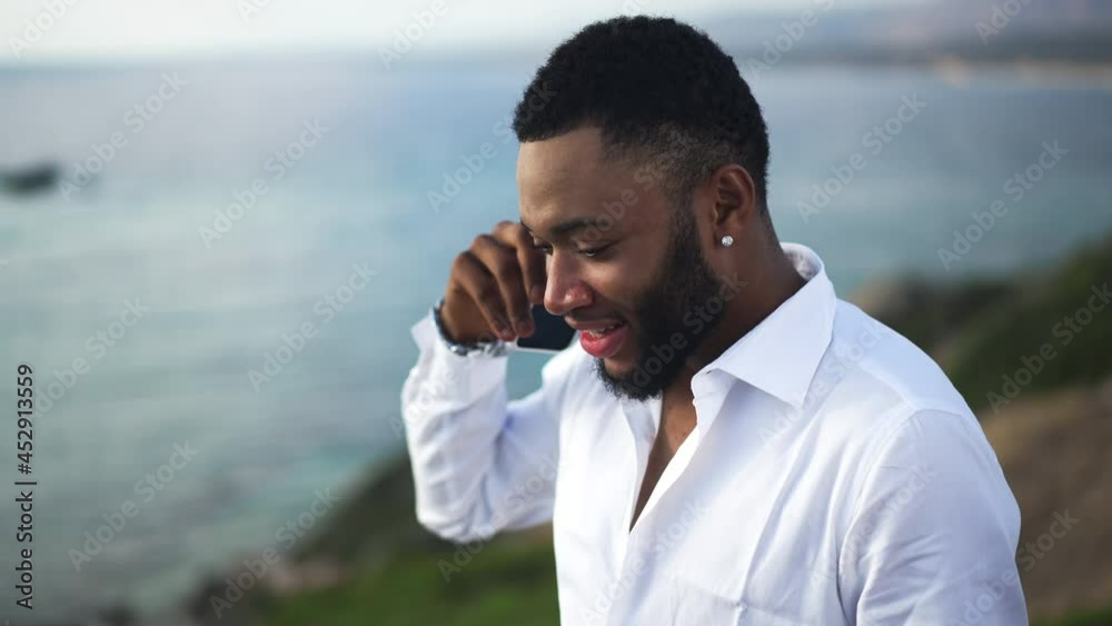 Confident handsome LGBT man talking on smartphone resting on Mediterranean sea resort. Portrait of smiling African American gay tourist at beautiful landscape with azure blue water Confident handsome LGBT man talking on smartphone resting on Mediterranean sea resort. Portrait of smiling African American gay tourist at beautiful landscape with azure blue water
