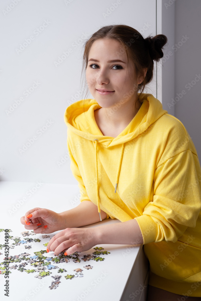 pretty brunette girl in yellow hoodie arranging pieces of a Jigsaw Puzzle Game in bright white room. playing board game home.