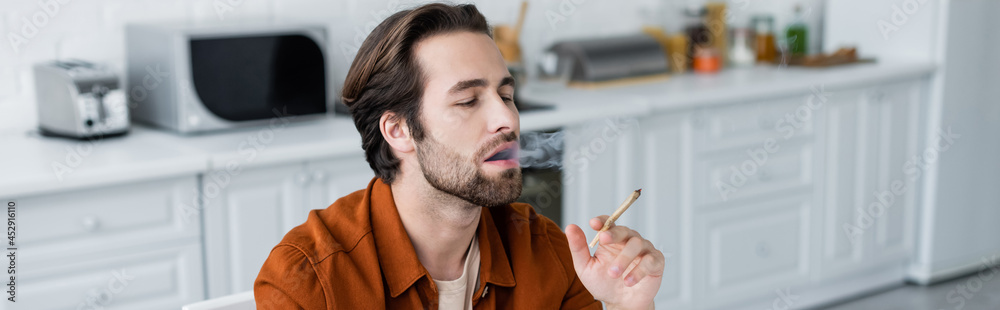 Obraz premium Man smoking joint of medical cannabis in kitchen, banner