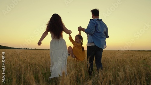 The child plays, jumps, runs next to mom, dad holding hands in the field. Happy family walk in a wheat field at sunset. A family of farmers. Parents, dear baby, happily walk together in rays of sun.