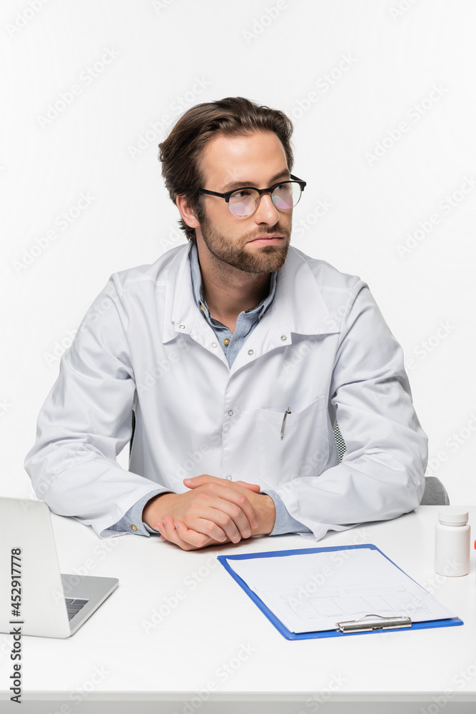 doctor sitting near clipboard, laptop and container with pills isolated on white, medical cannabis concept doctor sitting near clipboard, laptop and container with pills isolated on white, medical cannabis concept