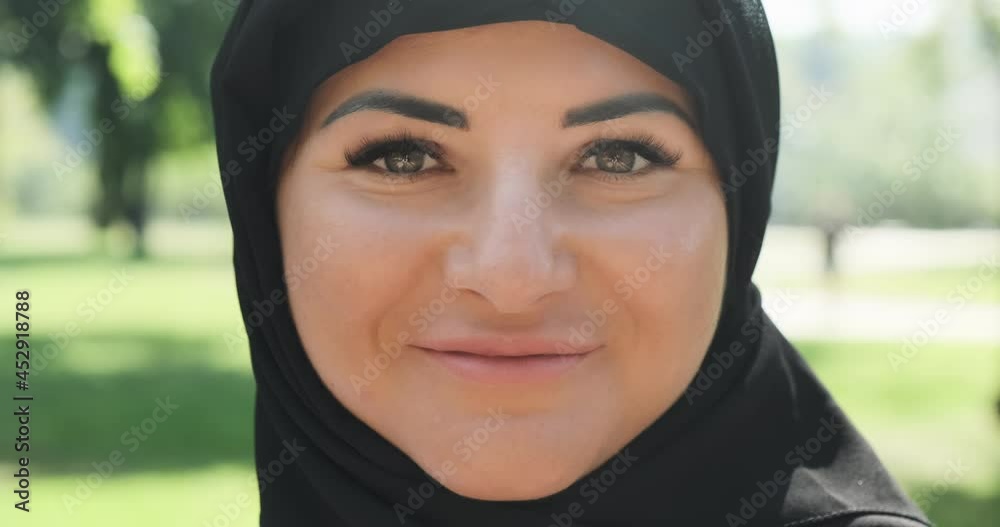 Close-up, muslim woman in hijab head scarf smiles. Beautiful muslim woman portrait, wearing black hijab head scarf in sunny summer park in morning, looking at camera. Confident islamic girl. Lifestyle