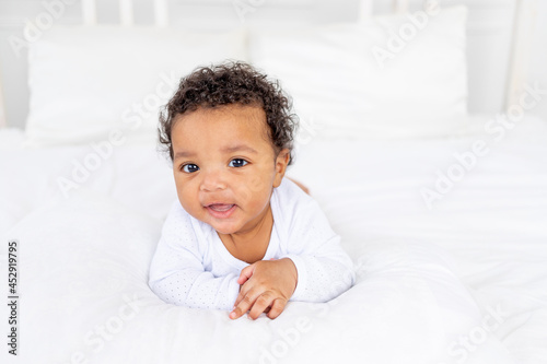 cute African-American little baby lying on a white bed in the bedroom of the house