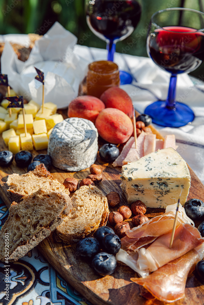 Cheese platter with different cow and goat cheeses, meat, nuts, Summer fruits and berries, jam and rye bread on wooden board.with red wine on little rustic table in the garden.