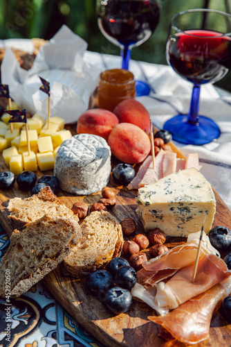 Cheese platter with different cow and goat cheeses, meat, nuts, Summer fruits and berries, jam and rye bread on wooden board.with red wine on little rustic table in the garden.