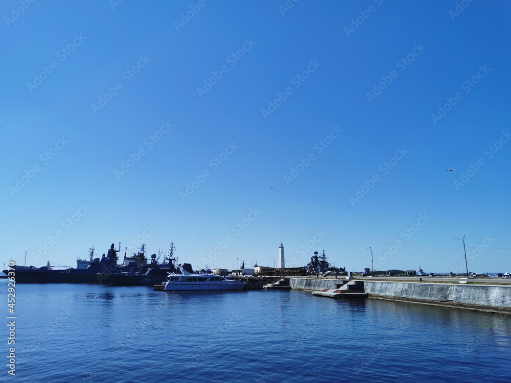 View of warships, boats and a pier with a lighthouse from Petrovskaya Embankment against the blue sky on a summer day in Kronstadt.