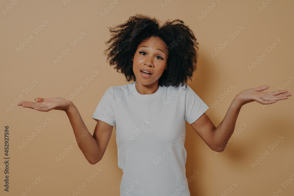 Unsure african woman woman shrugging shoulders and spreading arms sideways, isolated over beige wall Unsure african woman woman shrugging shoulders and spreading arms sideways, isolated over beige wall