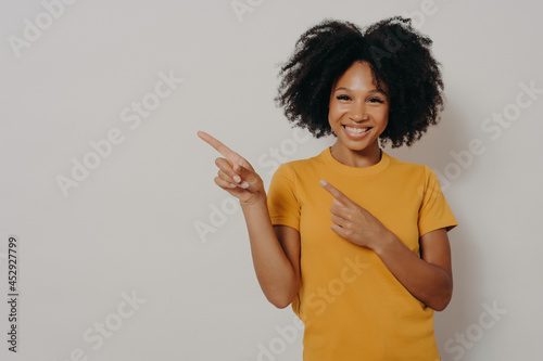 Cheerful dark skinned woman dressed in yellow tshirt pointing at left upper corner with forefingers