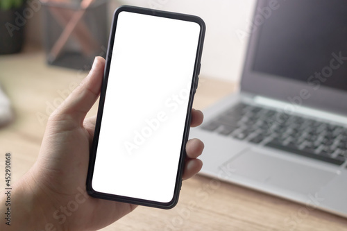 Man's hands holding or using smartphone with two hands in front of working space desk, laptop. Smartphone with white blank on screen, Cellphone mockup.