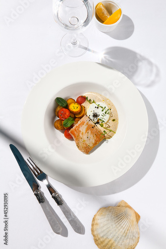 Fish steak with fillet tuna  warm tomato  baked potato and vitello tonnato sauce. Fish in white plate. Tuna sauce and vegetables. Contemporary seafood restaurant menu  summer day with sunlight shadow.