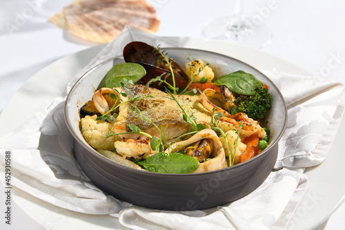 Elegant seafood stew with cod fish and vegetables in pot. Healthy fish dish - baked codfish, shrimp, mussels on broccoli and tomato. Serving white tablecloth and glass in summer day with shadow.