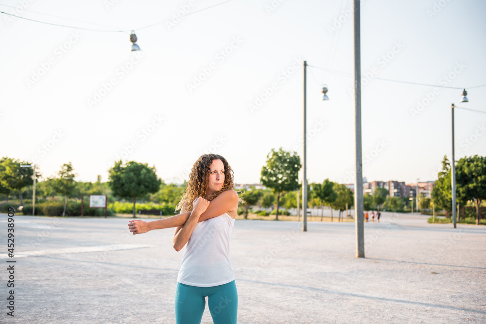 Caucasian curly-haired girl in a white t-shirt and blue leggings stretching her arms after a workout in a park, at sunset. active healthy lifestyle stretching Caucasian curly-haired girl in a white t-shirt and blue leggings stretching her arms after a workout in a park, at sunset. active healthy lifestyle stretching