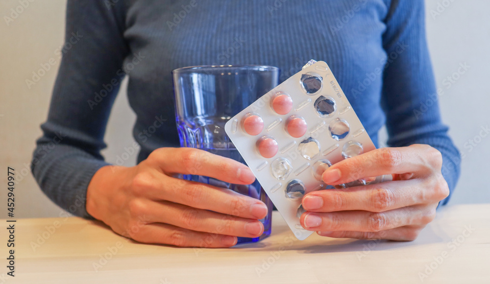 the girl is sitting at the table, holding pink pills in a package and a glass of water in her hands. Lack of vitamins in autumn and winter, uncontrolled medication the girl is sitting at the table, holding pink pills in a package and a glass of water in her hands. Lack of vitamins in autumn and winter, uncontrolled medication