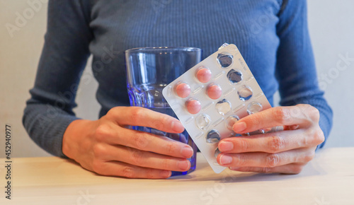 the girl is sitting at the table, holding pink pills in a package and a glass of water in her hands. Lack of vitamins in autumn and winter, uncontrolled medication