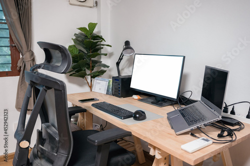 Wooden desk with laptop, display, mouse, lamp, ergonomic chair and artificial tree in workplace on bedroom