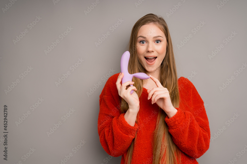 A pretty Caucasian girl in a red sweater depicts an emotion of surprise, opening her mouth and holding a lilac dildo, a massager for sex in her hand A pretty Caucasian girl in a red sweater depicts an emotion of surprise, opening her mouth and holding a lilac dildo, a massager for sex in her hand