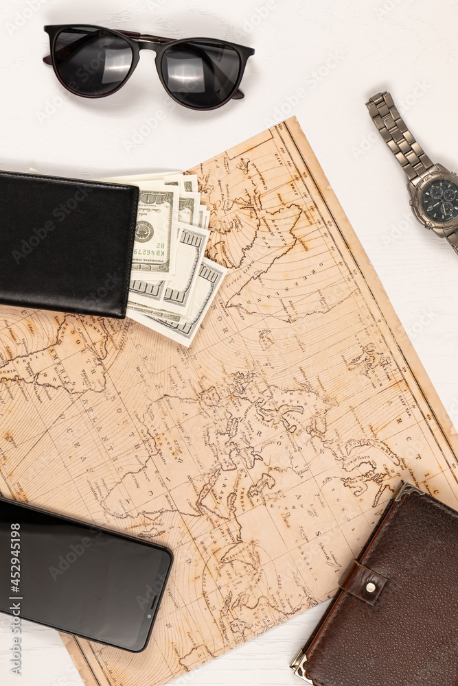 world map, smartphone, black leather wallet with money, diary, sunglasses and a watch on a white wooden table. view from above