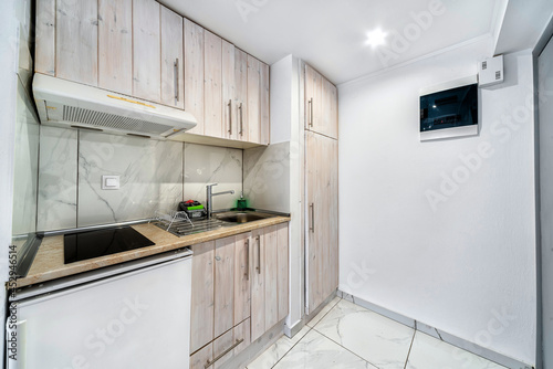 Modern kitchenette in light colours in apartment or studio or hotel room, equipped with mini frigde, small ceramic cooker and hood, sink and many diffrenet lockers cabinets.  photo