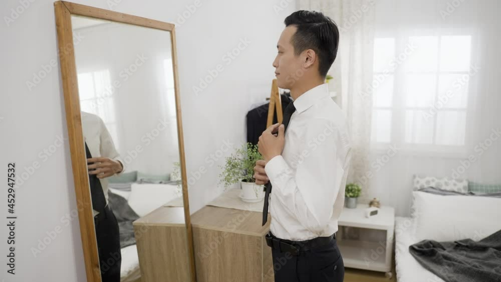 confident asian business man looking at himself in the mirror with folded arms while adjusting tie and hair preparing for meeting in the bedroom at home confident asian business man looking at himself in the mirror with folded arms while adjusting tie and hair preparing for meeting in the bedroom at home