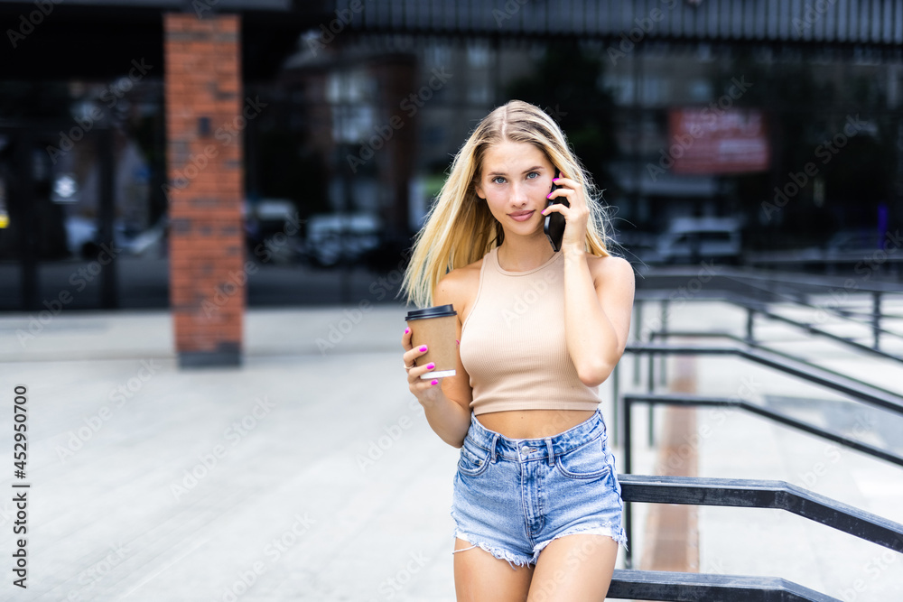 Fototapeta premium Young woman with coffee cup talking on the phone and walking at the city street