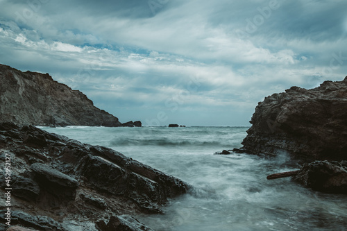 rocky beach landscape during storm with cloudy sky. Dramataic long exposure of sea waves breaking on rocks.