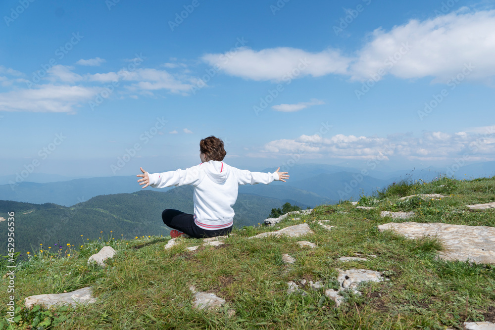 Young female with arms outstretched is sitting on top of mountain peak with sense of freedom. Copy space, healthy life and relaxation concept