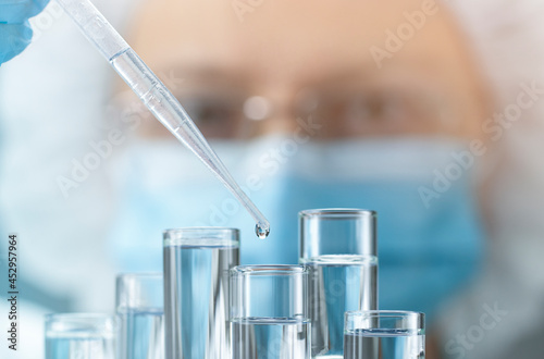 Scientist or assistant in protective mask adds liquid from pipette to one of test tubes. Concept of science development, genetic research, pharmaceutical technology or biotech science.