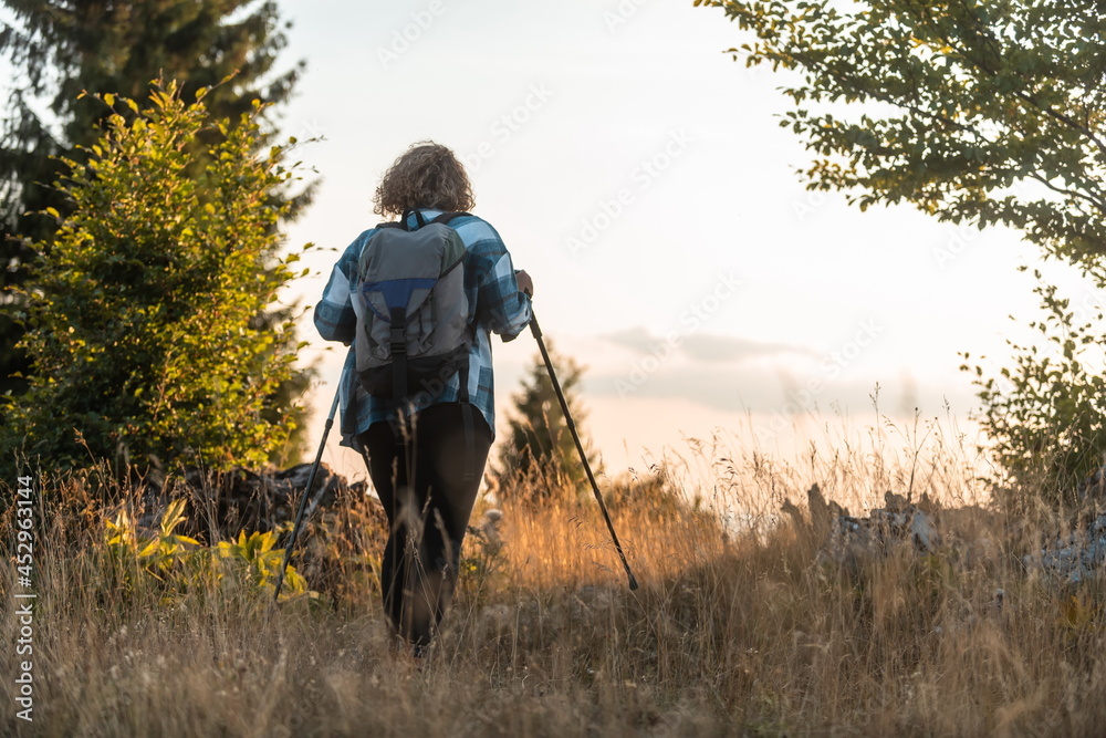 A woman with sticks, a backpack on her back, and mountaineering equipment walking on top of a mountain at sunset.
