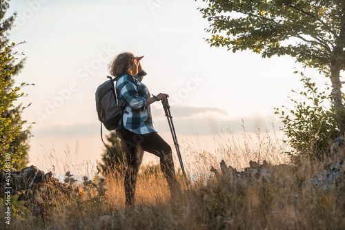 A woman with sticks, a backpack on her back, and mountaineering equipment walking on top of a mountain at sunset. © Minet