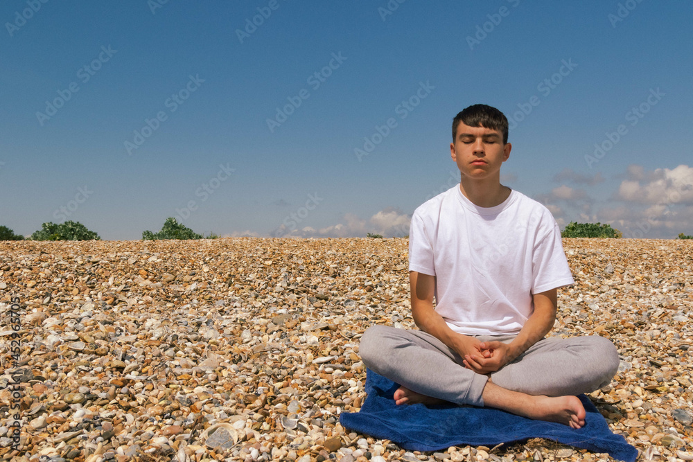 A Caucasian teenage boy meditating on a stony beach with his hands resting on his lap
