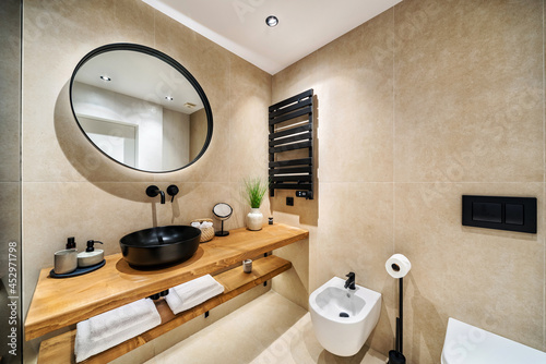 Modern tiled bathroom in beige and brown warm colors.  It has  wooden stand for black sink  bidet and round mirror on the wall. 