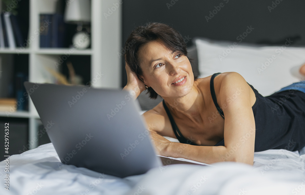 Caucasian woman in casual wear lying on comfy bed with wireless laptop. Middle aged lady with brown hair looking aside and dreaming about something. Caucasian woman in casual wear lying on comfy bed with wireless laptop. Middle aged lady with brown hair looking aside and dreaming about something.