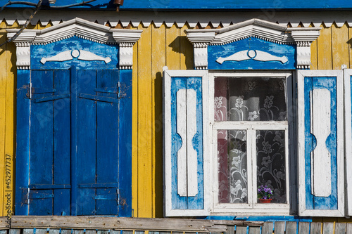 The bright colorful facade of a wooden village house in yellow with carved blue shutters on the windows.