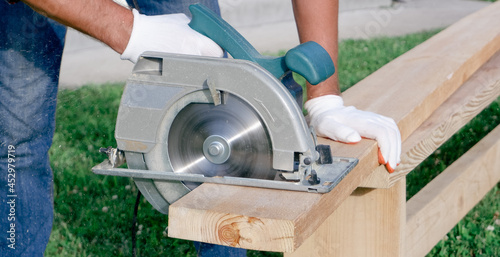 A male joiner works with a hand-held circular saw. A builder is sawing a board at the construction site of a new house.