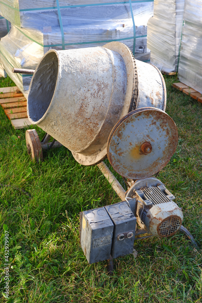Industrial concrete mixer at a construction site. Dirty old concrete mixer on the grass against the background of pellets with building materials Industrial concrete mixer at a construction site. Dirty old concrete mixer on the grass against the background of pellets with building materials
